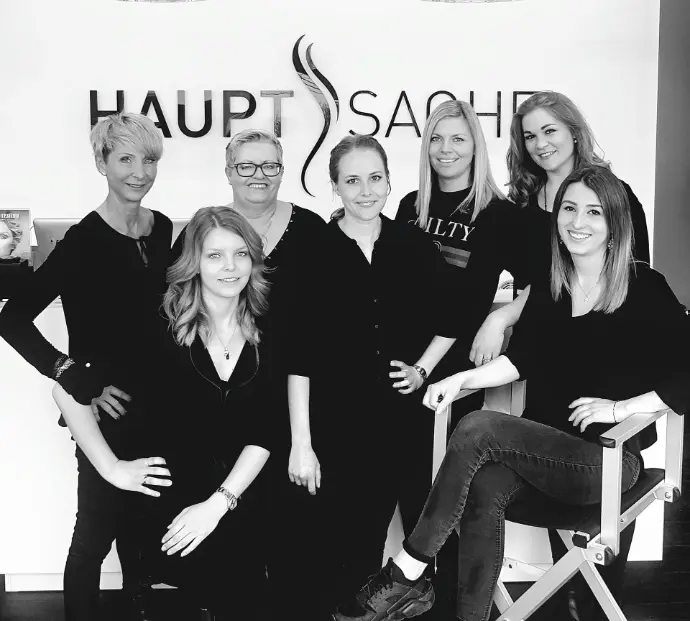 Schwarz-weiß Gruppenfoto des Teams von Coiffeur HAUPTSACHE Sursee vor dem Salon-Logo, freundlich und professionell